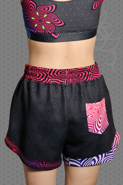 back view psychedelic loose-fit shorts with black geometric pattern body, magenta-to-purple optical pattern panels and waistband, gradient pink-purple-blue accents, drawstring waist, and contrasting patterned back pocket
