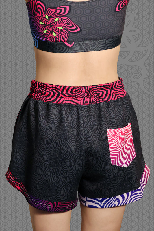 back view psychedelic loose-fit shorts with black geometric pattern body, magenta-to-purple optical pattern panels and waistband, gradient pink-purple-blue accents, drawstring waist, and contrasting patterned back pocket